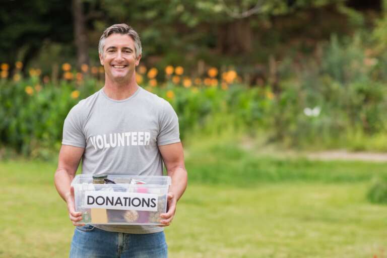 Man standing on lawn wearing VOLUNTEER Tshirt holding DONATIONS bin containing food items